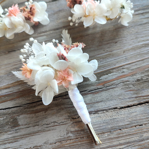 Boutonnière florale Clara avec des fleurs éternelles blanches et roses, posée sur un fond en bois naturel.