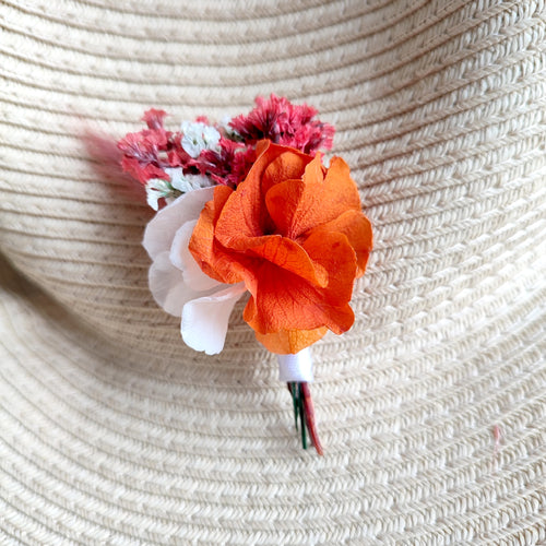 Broche florale Léna épinglée sur un chapeau en paille, avec des fleurs stabilisées orange, blanc et rouge.